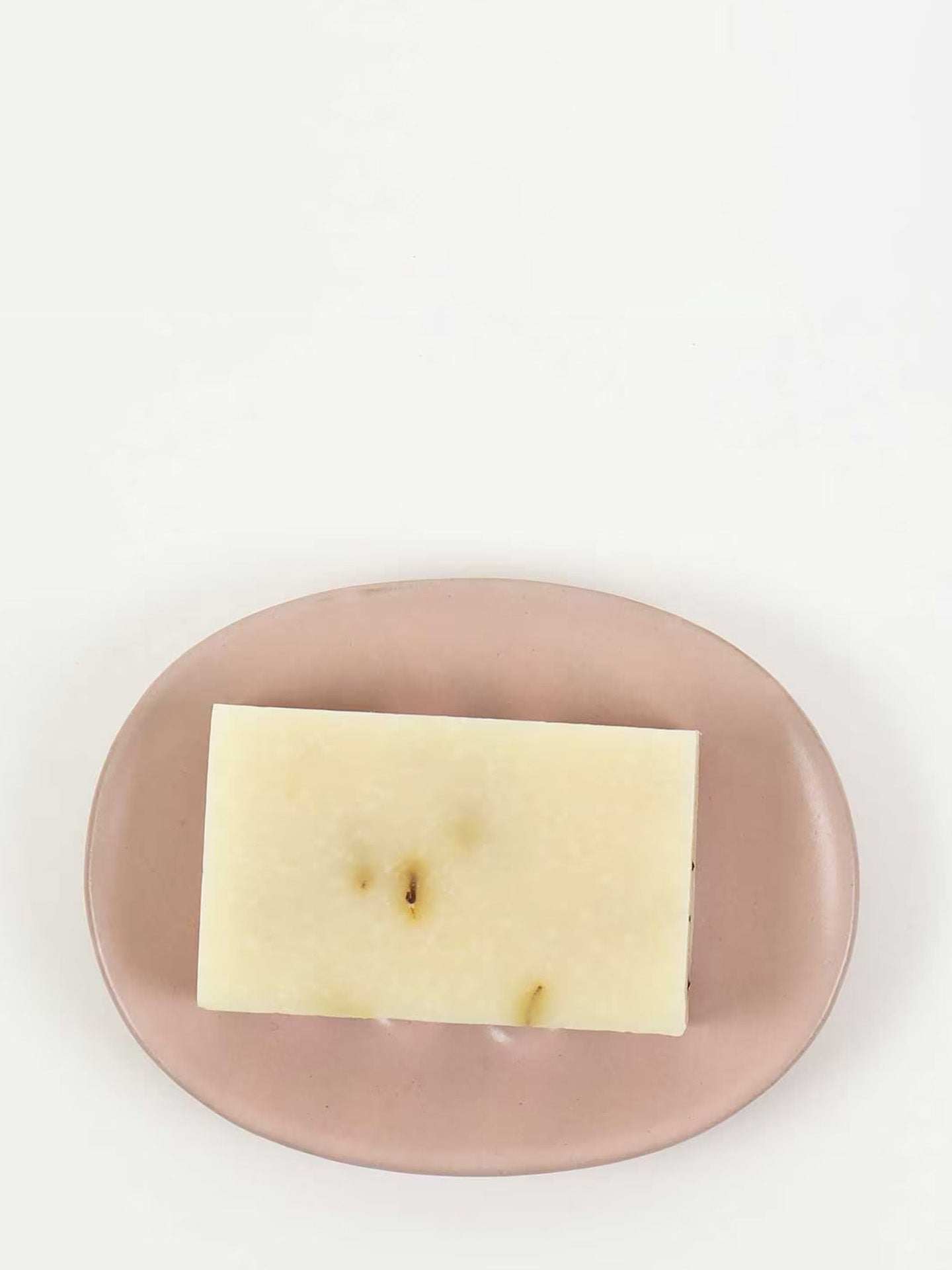 A rectangular bar of pale cream soap with small brown specks, displayed on a dusty pink oval soap dish against a clean white background.