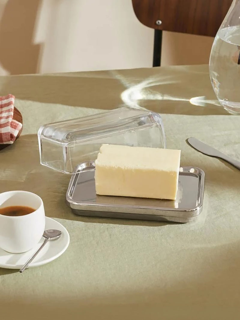 A rectangular metal butter dish with a clear glass lid, holding a stick of pale yellow butter, set on a beige tablecloth with a cup of coffee, a butter knife, and a teak chair in the background.