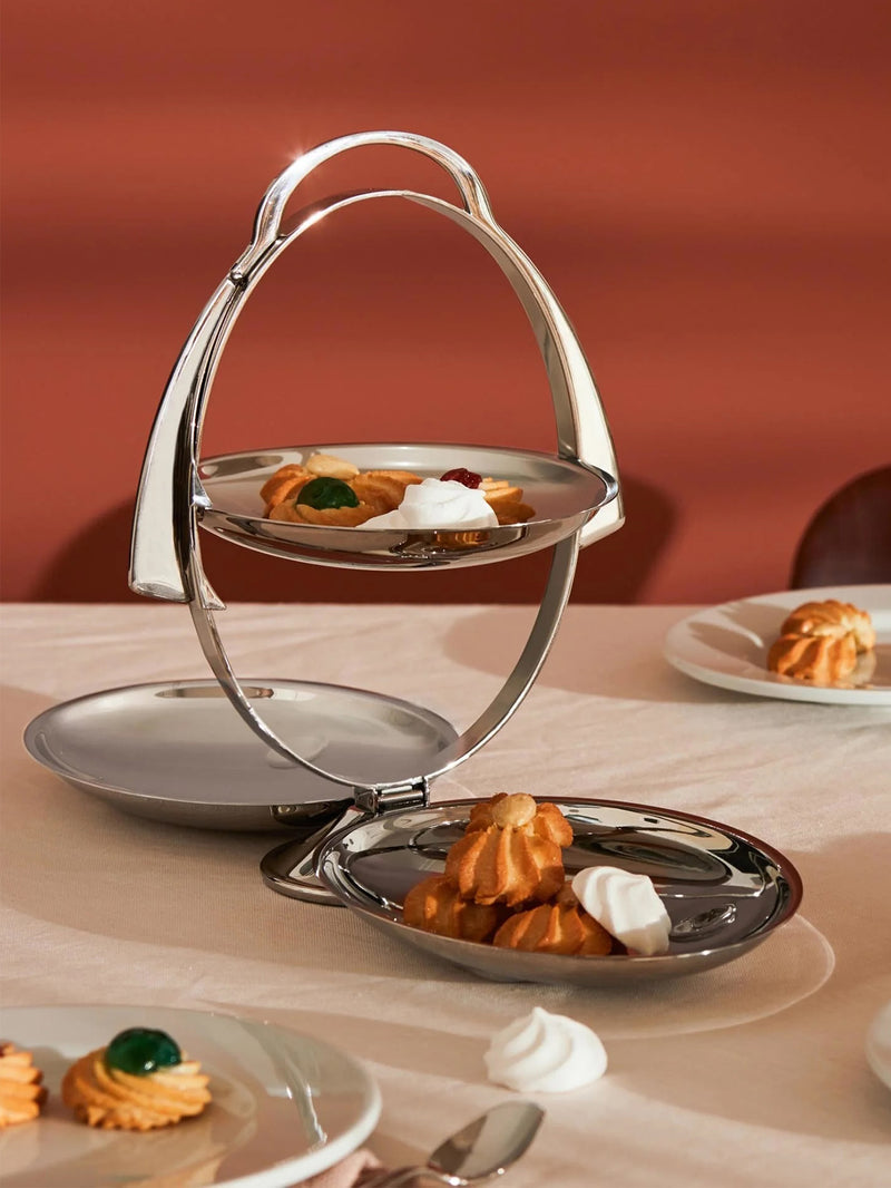 A polished silver metal two-tiered serving stand with three serving plates with one on top, holding assorted pastries and meringues, set on a table with other treats and plates, against a warm terracotta background.