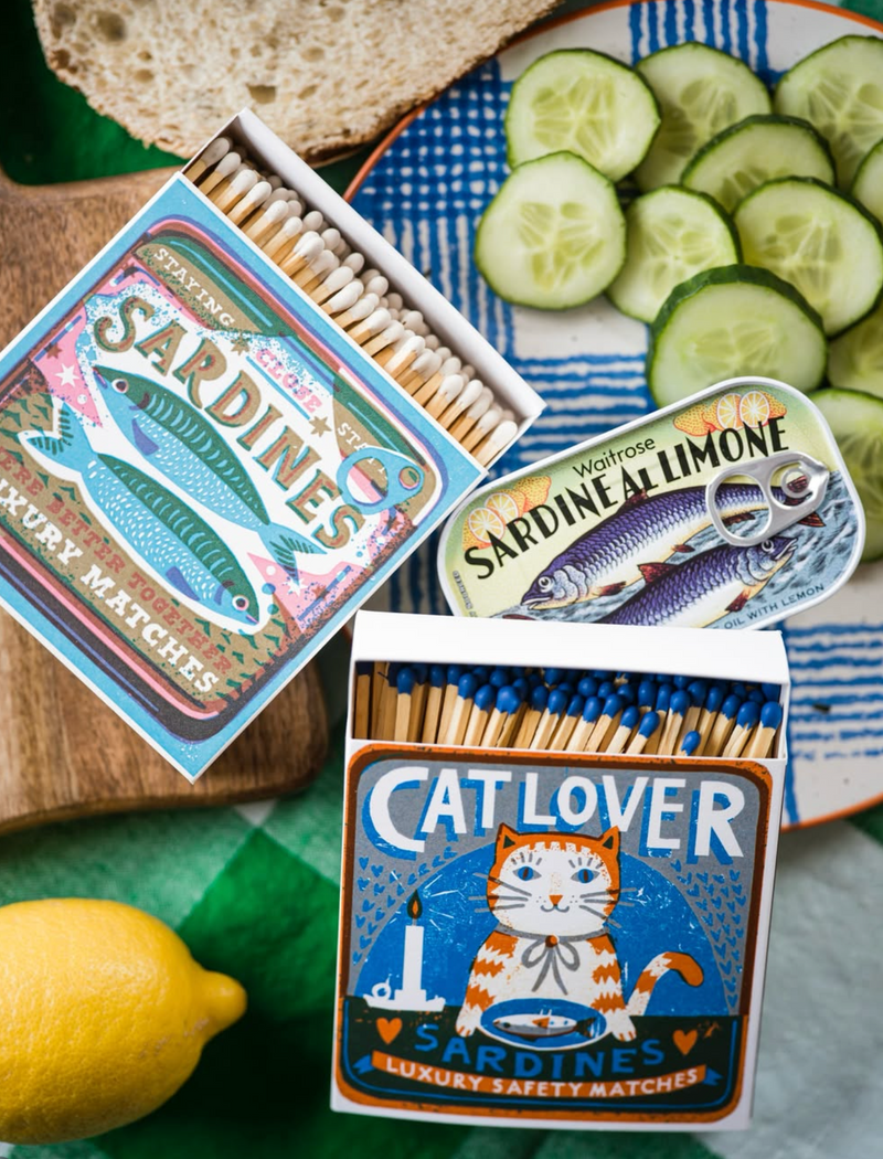 Three matchboxes with different designs on a table with sliced cucumbers and a lemon.