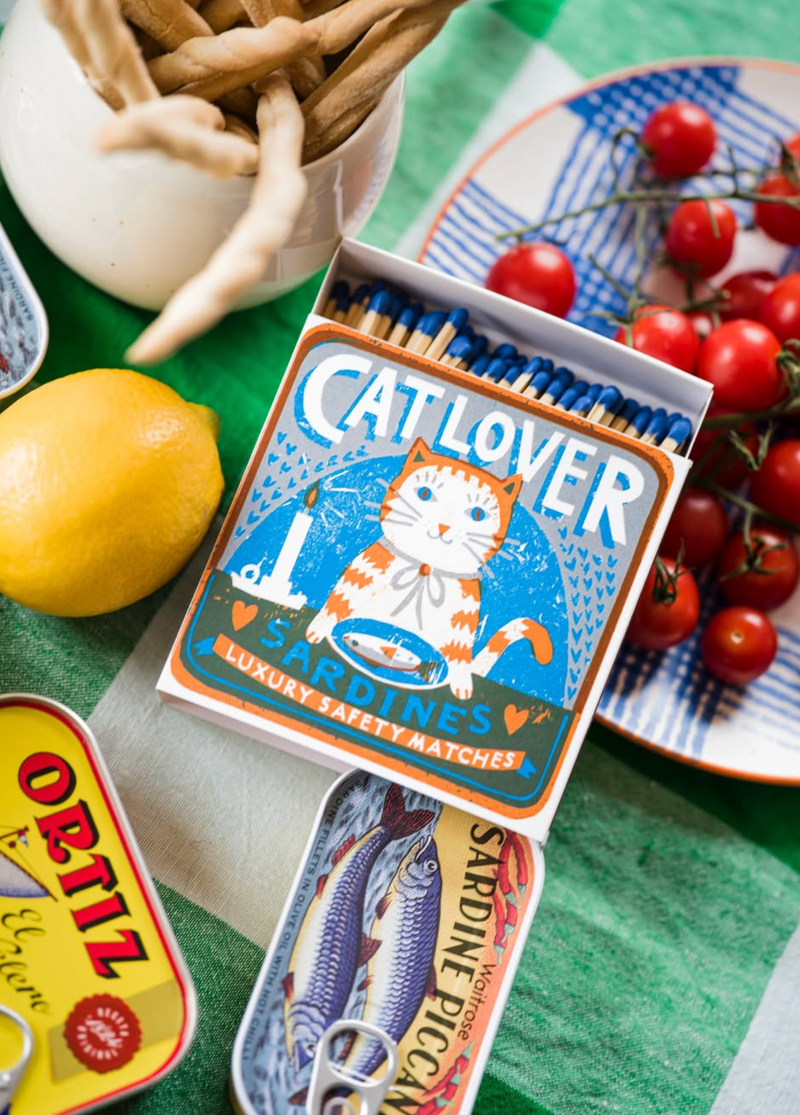 Matchbox labeled 'Cat Lover' with a cat illustration on a table with tomatoes and a lemon.