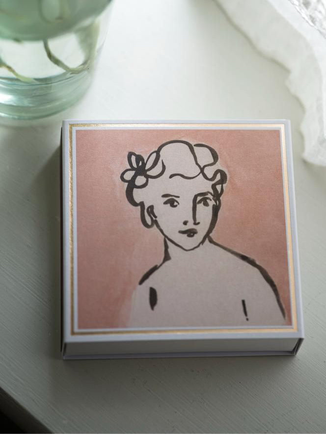 Matchbox with a stylized illustration of a woman on a pink background, placed on a white surface.