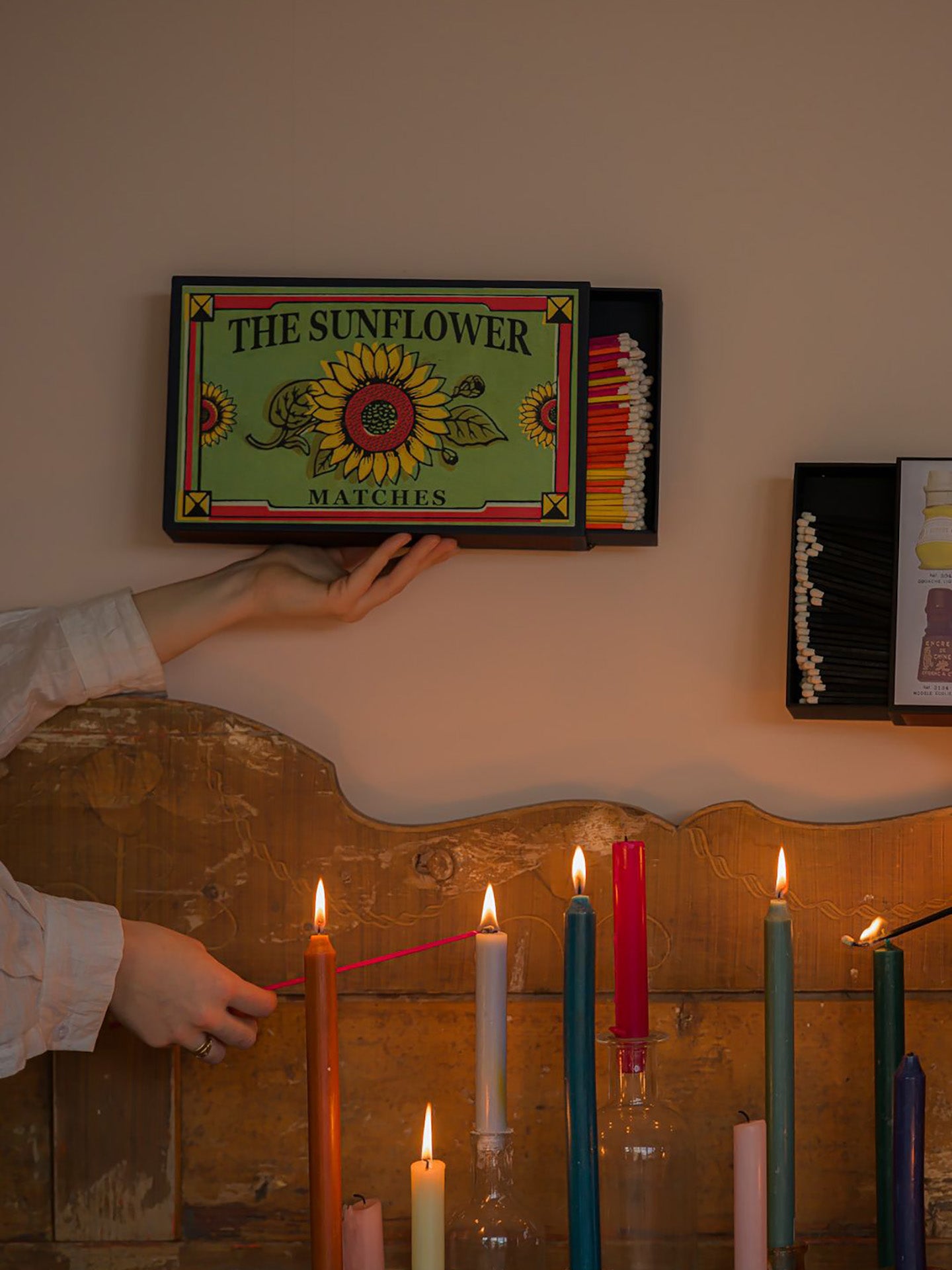 A person holds an unusually large vintage-style matchbox labeled 'THE SUNFLOWER MATCHES' and on the other hand is lightng a candle with a very big match stick in front of an old wooden sofa back against a beige wall above a with lit candles.