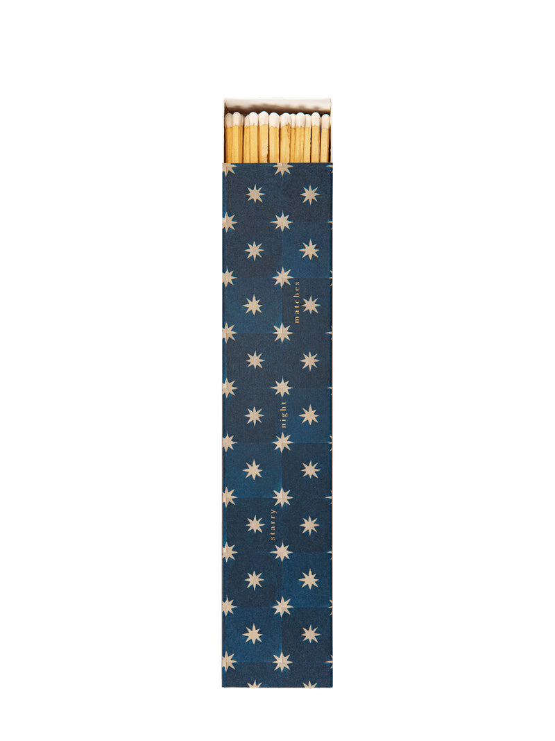 A long and thin match box with illustrated cover of geometric star pattern depicting a dark blue sky and white stars and the text starry night matches printed with small golden letters and the white-tipped match sticks visible from the top end, photographed against a white background