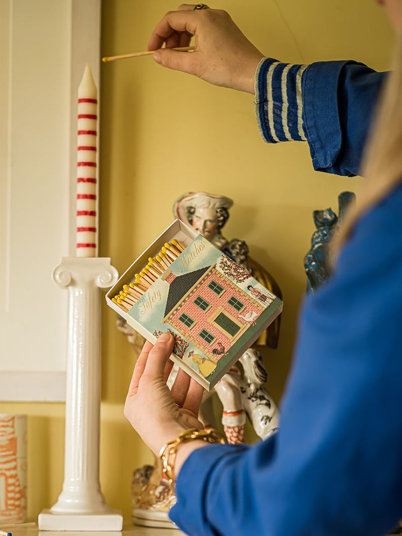 A person in a blue jacket holds an open decorative matchbox with a colorful house illustration and yellow-tipped matches, while another hand is holding an unlit match next to a tall white candle with red stripes in a warm indoor setting.