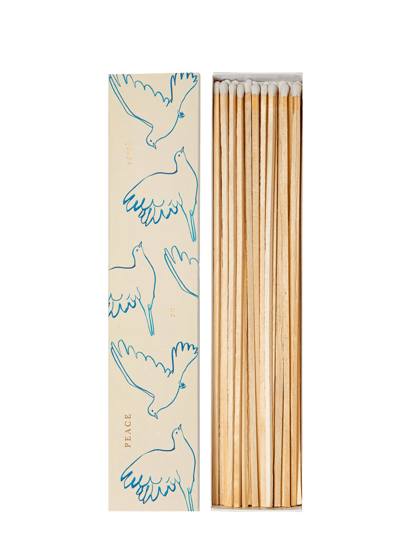 A set of wooden matchsticks in an open rectangular box next to its cream-colored lid, which features blue line drawings of flying doves and the words 'PEACE ON EARTH' printed vertically.