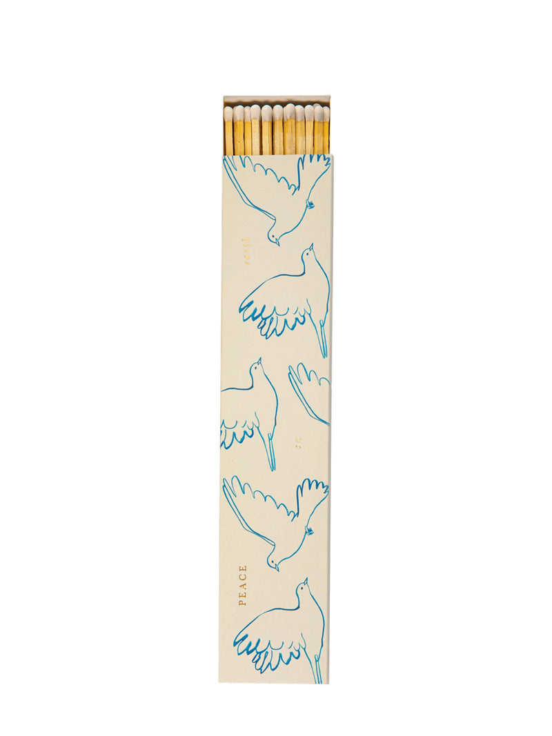 A little opened tall rectangular matchbox with a cream-colored cover printed with blue line drawings of flying doves and the words 'PEACE', 'ON' and 'EARTH' in gold, white-tipped matches visible at the top, and the box is photographed on a plain white background.