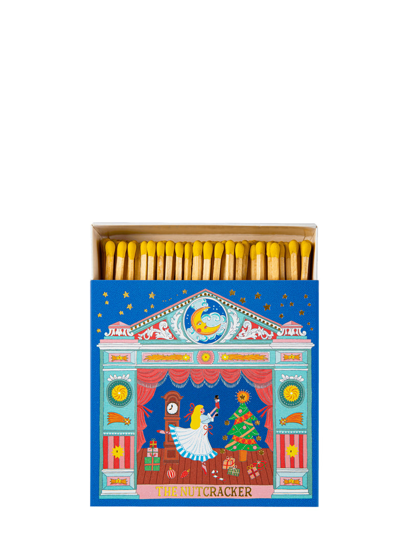 A decorative matchbox filled with yellow-tipped matches, shown open against a plain white background. The matchbox lid features a colorful illustrated scene with a clock, a nutcracker, stars, and a blue background.