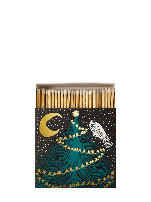 A matchbox with a festive Christmas illustration, featuring a teal tree, gold crescent moon and garland, and a white bird on a black background. The box is open to reveal wooden matchsticks with white tips.