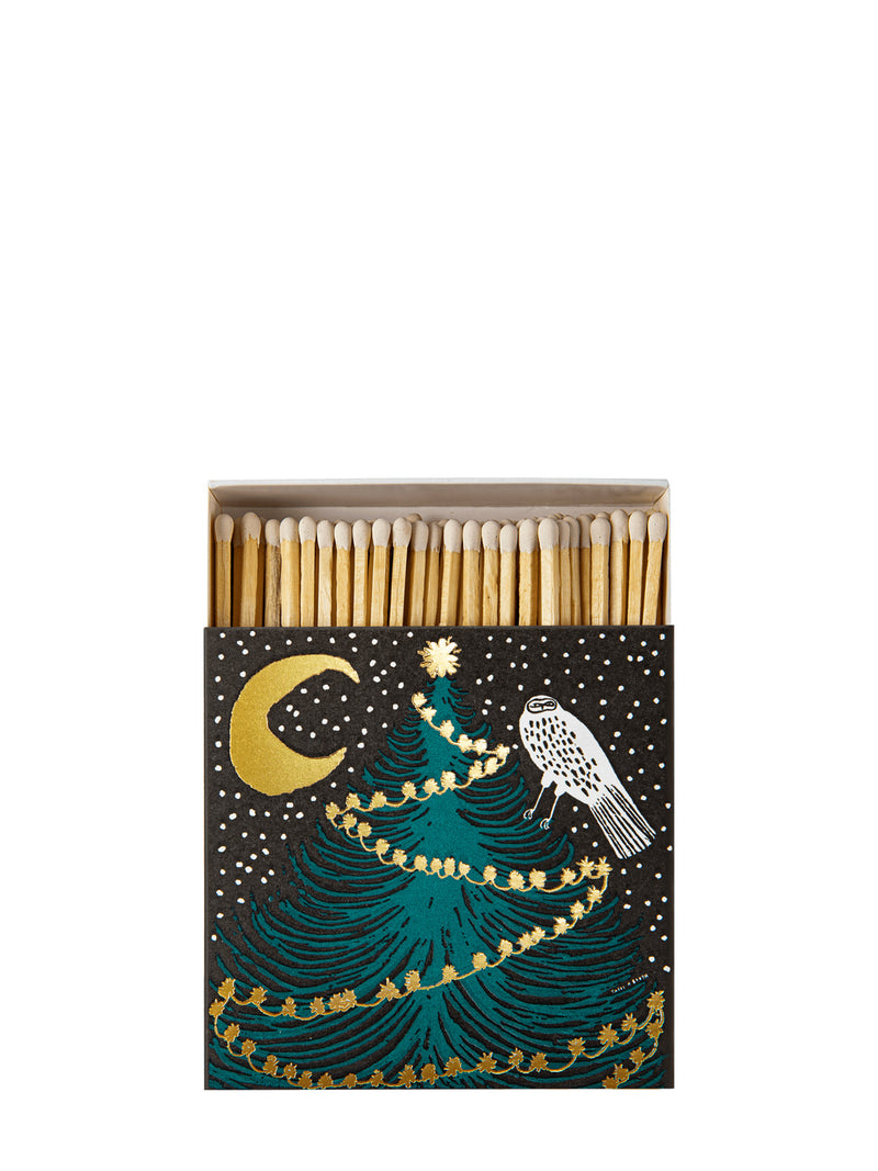 A matchbox with a festive Christmas illustration, featuring a teal tree, gold crescent moon and garland, and a white bird on a black background. The box is open to reveal wooden matchsticks with white tips.