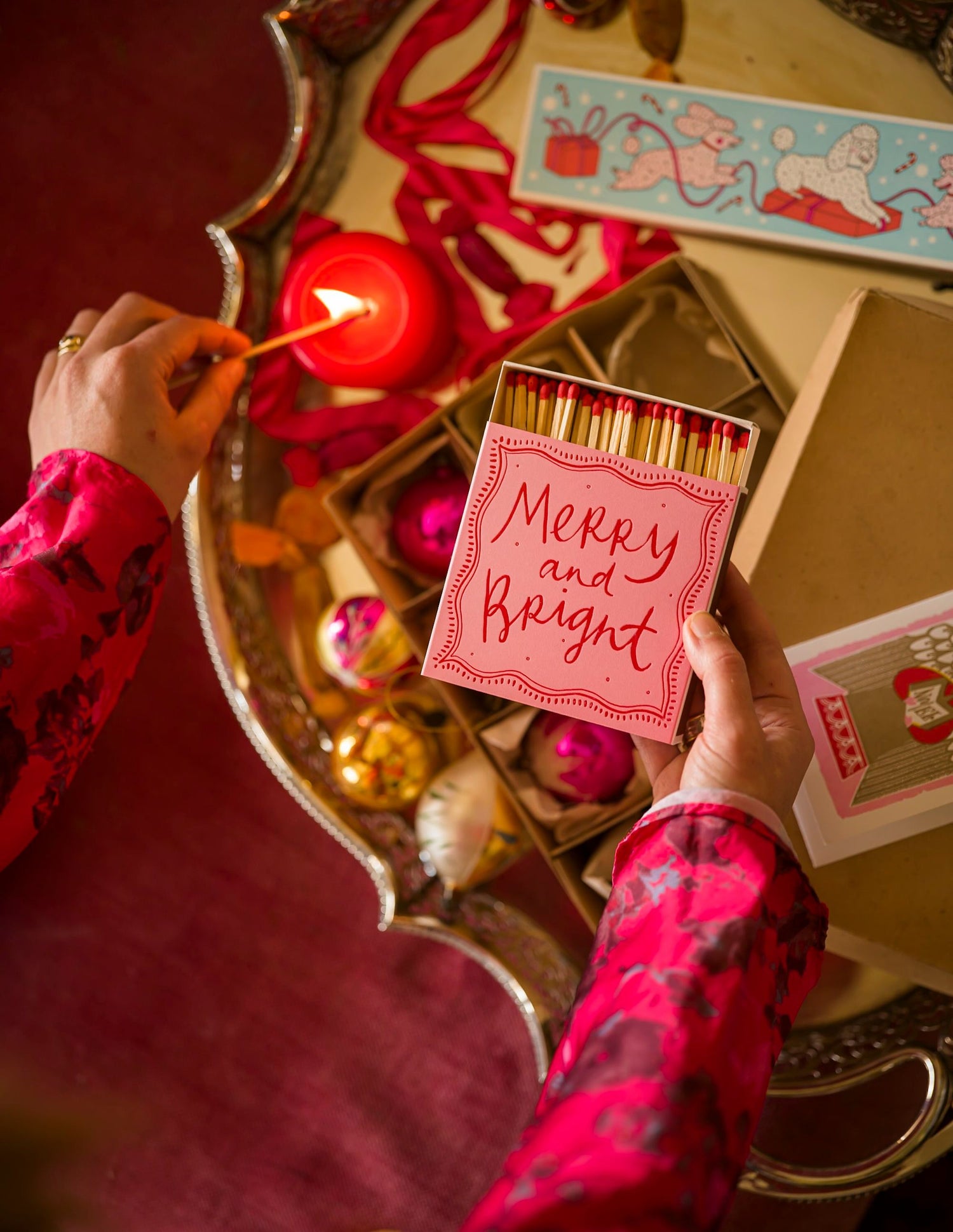 Person lighting a candle with a box of matches labeled 'Merry and Bright' on a decorative table.