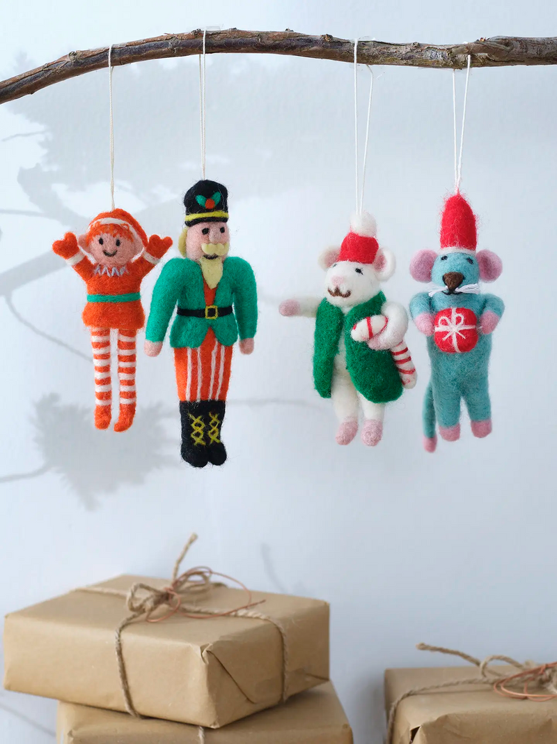 Four handmade felt Christmas ornaments hanging from a rustic wooden branch against a light blue wall over some Christmas presents wrapped in kraft paper and natural string bows. The ornaments are all smiling and include an elf in orange and white, the Nutcracker from the Nutcracker ballet in green and red, a white mouse in green vest and red hat holding a candy cane, and a blue mouse in red Christmas elf hat, holding a present with white whiskers.