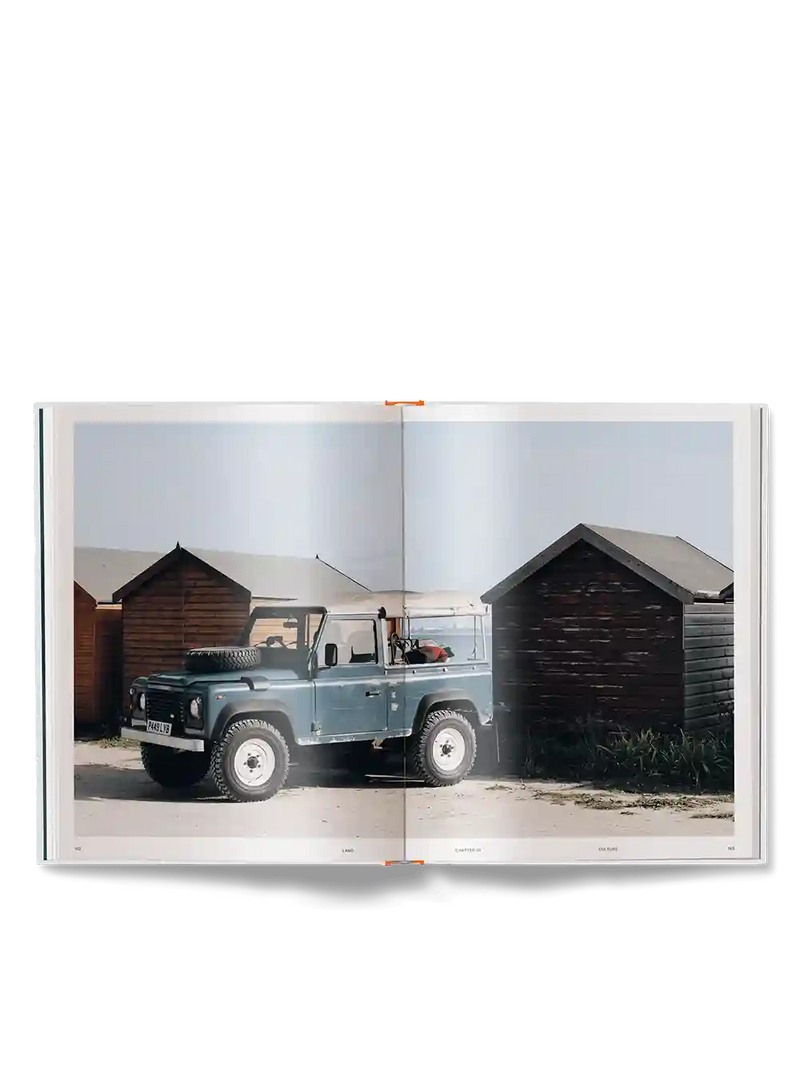 An open book or magazine spread showing a vintage blue off-road vehicle parked between wooden sheds under a pale sky.