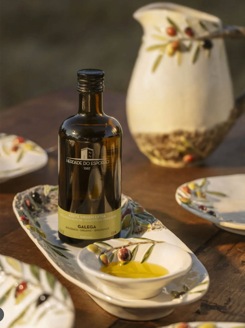 Bottle of olive oil on a wooden table with a decorative pitcher and plates in the background