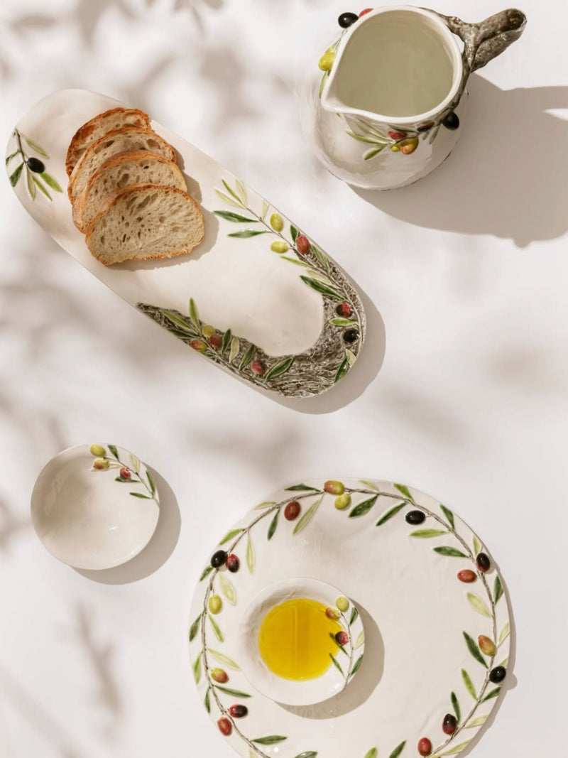Ceramic plates with olive design, bread, and a pitcher on a light surface.