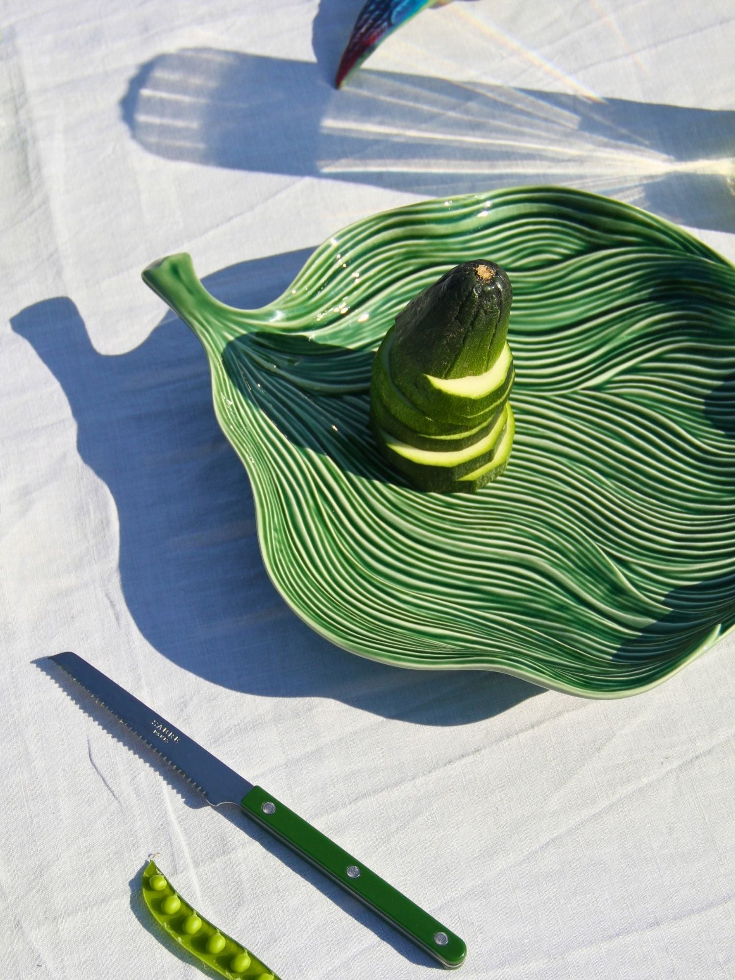 A green ceramic leaf-shaped serving dish with ridged detailing, styled with a zucchini and a pea pod on a white fabric background, alongside a matching green-handled knife.