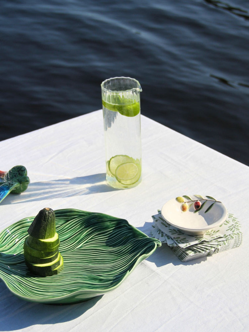 A styled outdoor table setting with a tall clear glass pitcher containing lime slices, a green leaf-shaped plate holding a cucumber, and a small white dish on a folded napkin, all set against dark blue water.