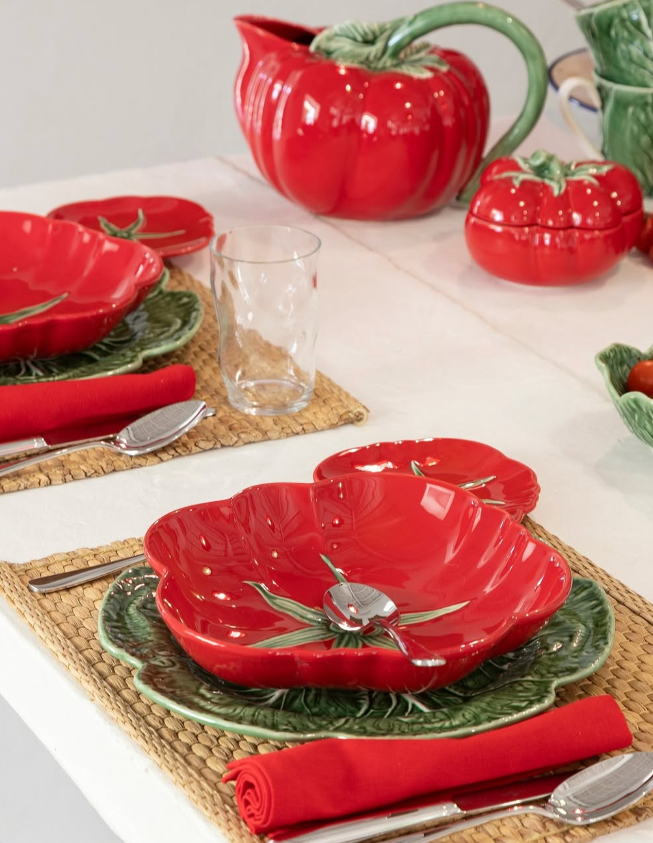 Red ceramic tomato-shaped bowls and pitcher by Bordallo Pinheiro on a white table with red napkins and silverware.