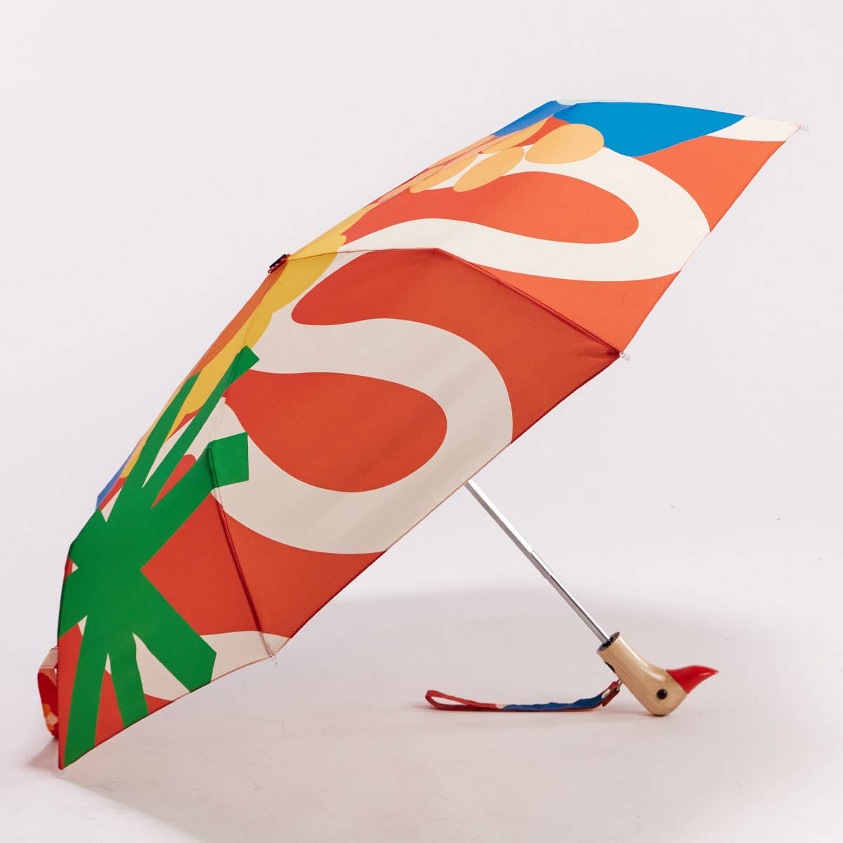 A folding umbrella with a bold retro abstract pattern in orange, white, green, yellow, and blue, featuring a light wood handle shaped like a duck's head with a red beak, and a small red-and-blue wrist strap, photographed on a clean white background.