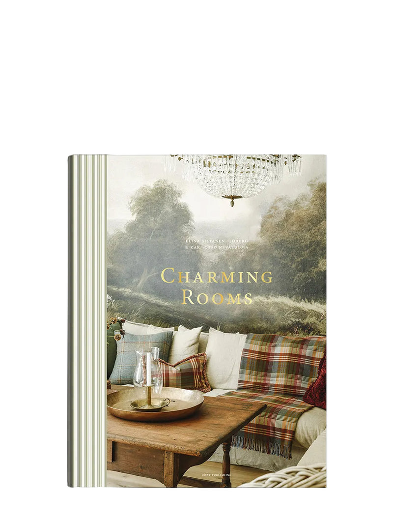 A hardcover book titled 'Charming Rooms' by Martha Stewart, featuring a cover photo of a cozy living room with a plaid sofa, wooden coffee table, and a misty forest background.