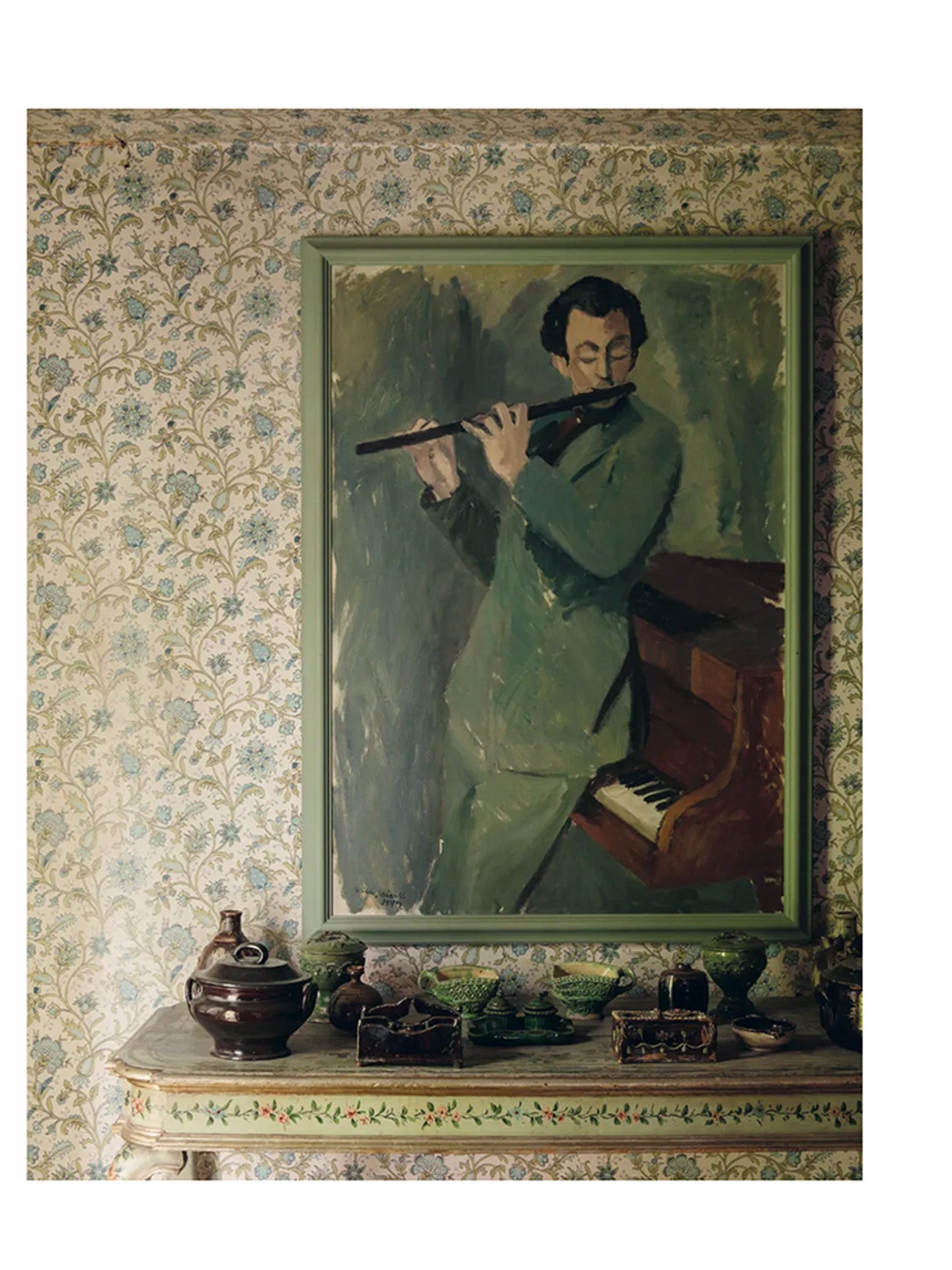 Page from the book Nordic Atelier Homes showing soft beige and sage green interior with patterned wallpaper and framed painting of a man playing a flute hung above of a little side table with floral and leaf decoration and various ceramic dishes and candleholders on the table.