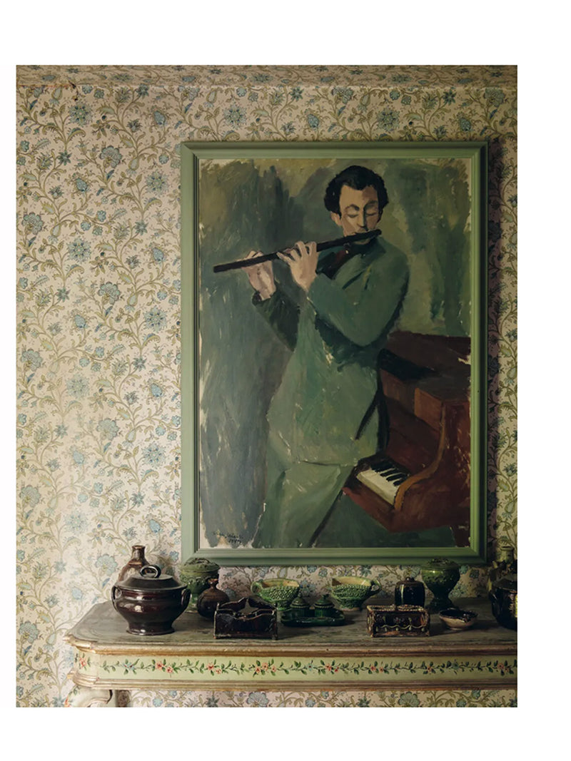 Page from the book Nordic Atelier Homes showing soft beige and sage green interior with patterned wallpaper and framed painting of a man playing a flute hung above of a little side table with floral and leaf decoration and various ceramic dishes and candleholders on the table.
