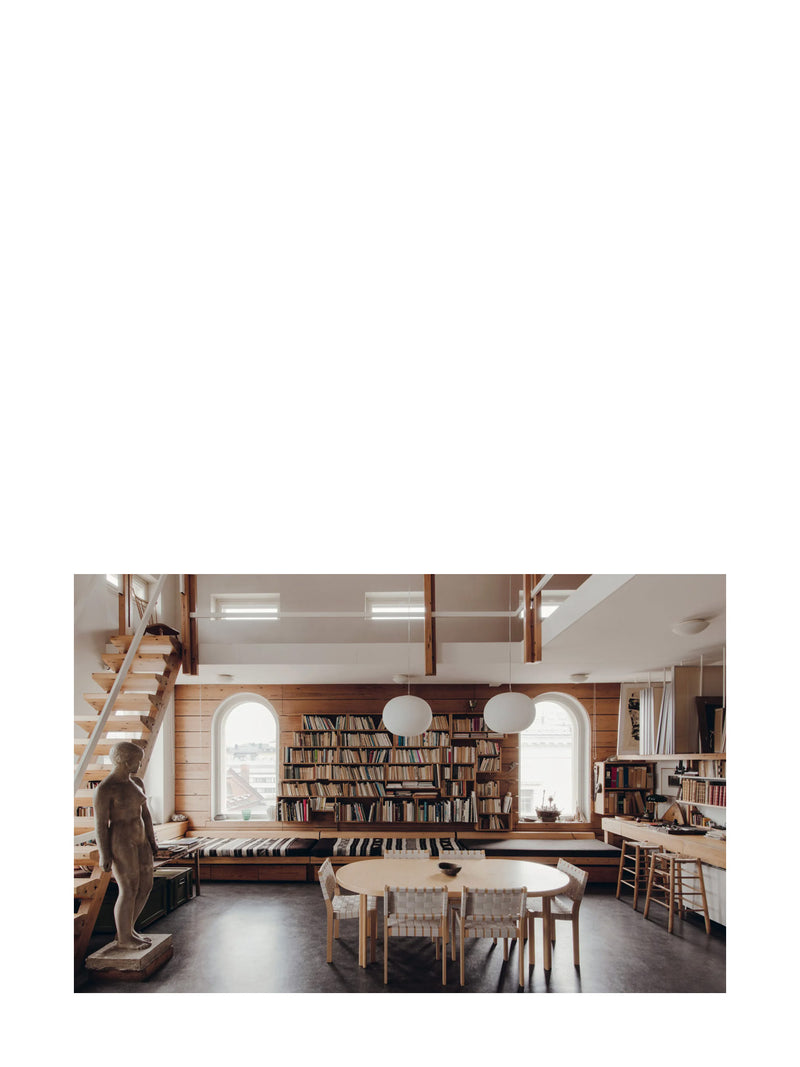 Atelier interior design of Tove Jansson's studio home with a arched windows, bookshelf, Artek dinner table, and chairs and stairs leading up the mezzanine next to a life-sized realistic sculpture of a woman standing.