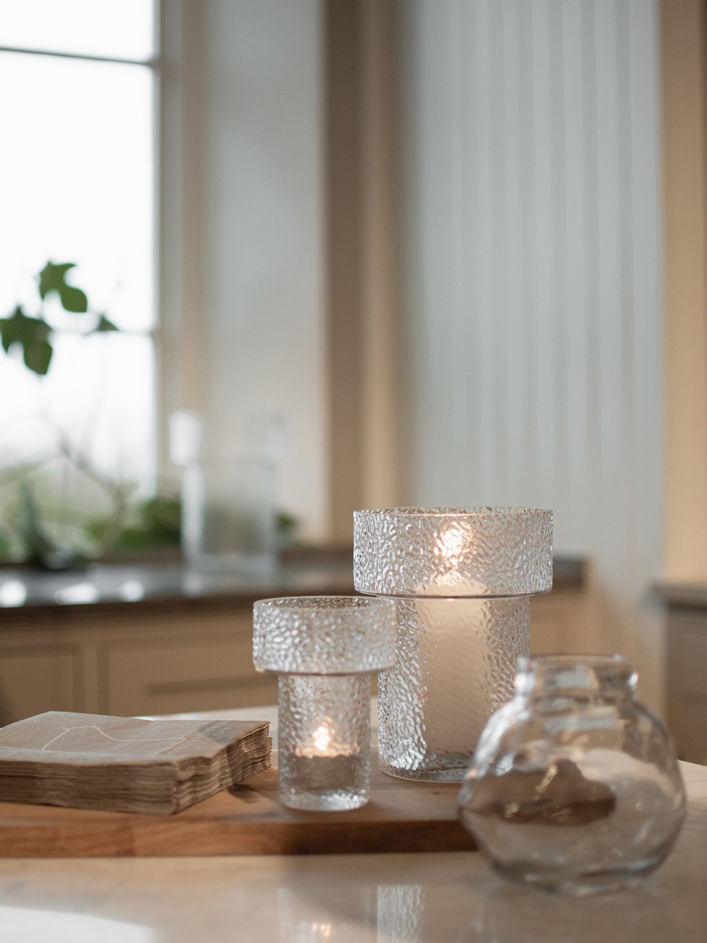 Two textured glass vases acting as candle holders with lit tea light on the smaller and a white table candle on the larger one, placed together on a wooden tray, alongside a clear glass vase, set in a softly blurred kitchen environment.