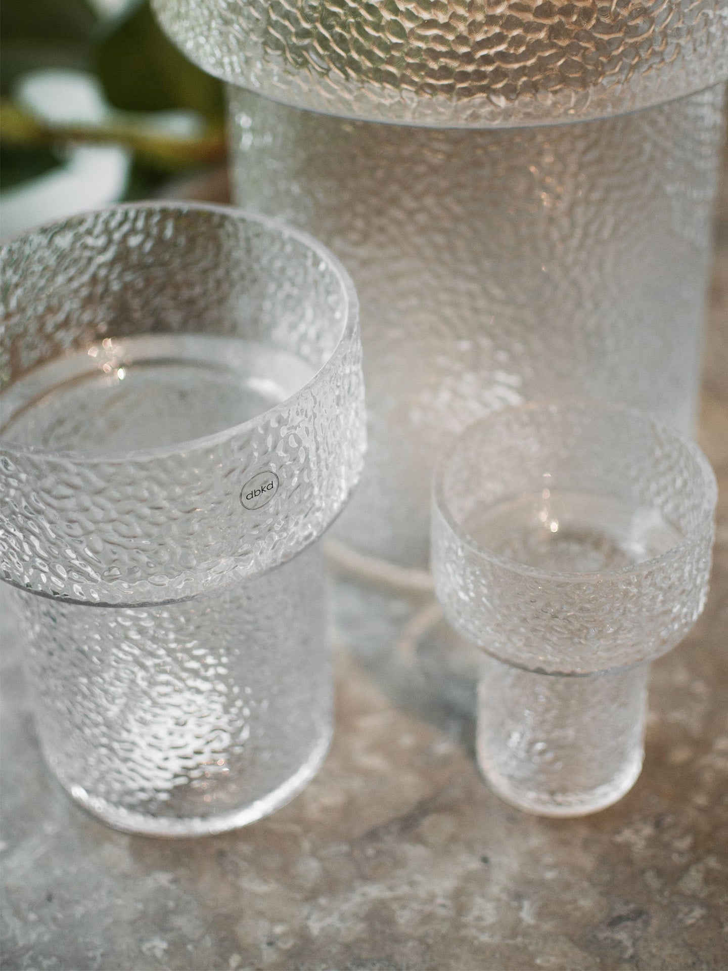 A set of clear textured glass vases, including a large, medium and a small Keeper structured vase, featuring a hammered or dimpled finish, placed on a stone-like surface.