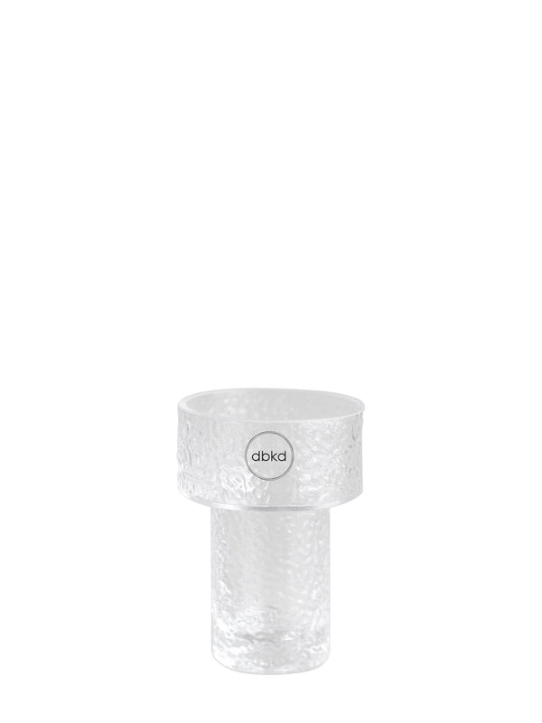 A clear textured glass flowers vase with a wide top rim and a cylindrical base, featuring a circular logo sticker with the brand name 'dbkd' in the center.
