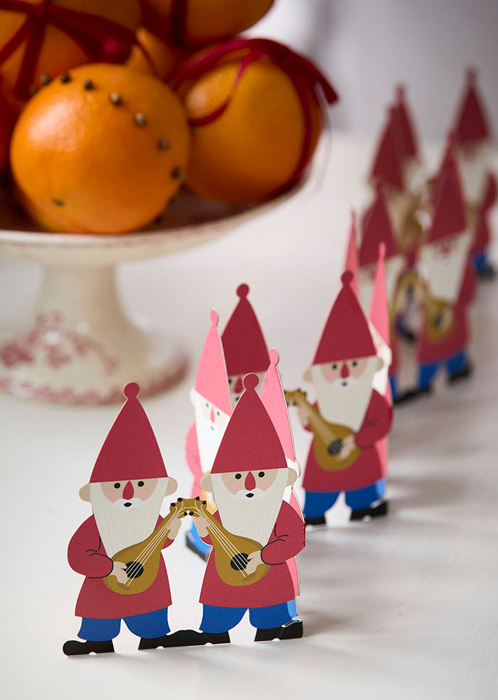Decorative paper gnomes with red hats and blue pants on a white surface, with oranges in the background.