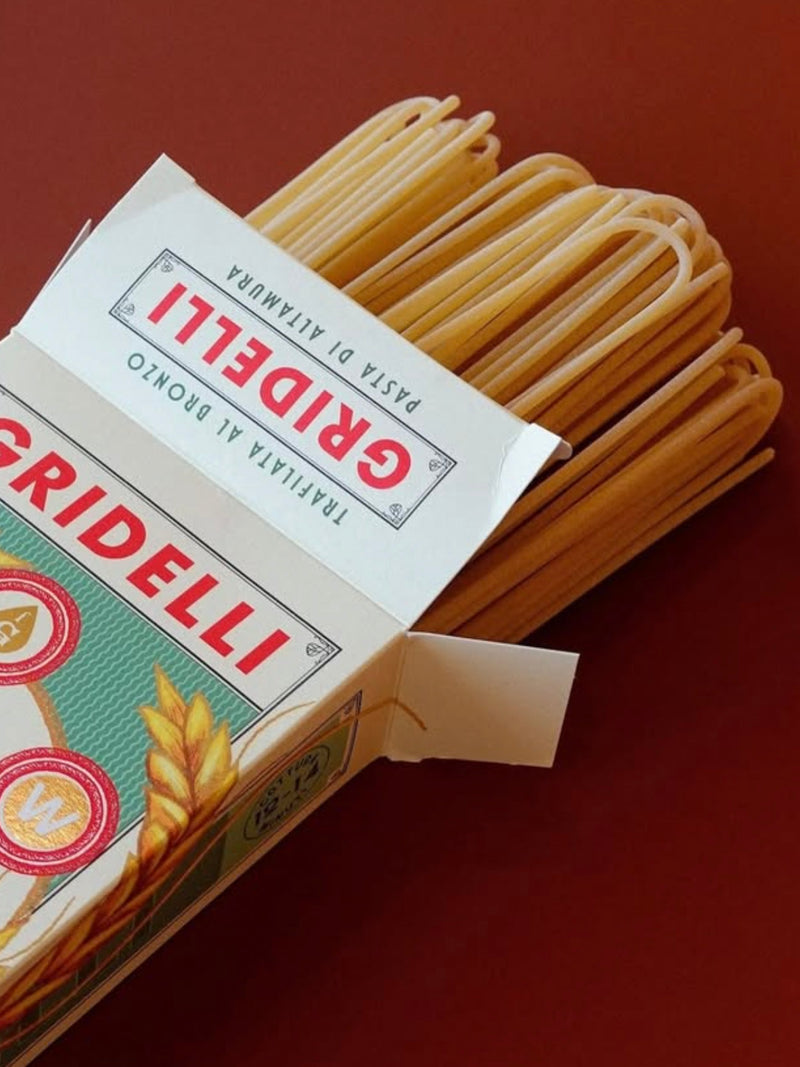 An open box of Gridelli spaghetti pasta on a solid dark red background, with the pale yellow pasta strands visible extending out of the packaging.