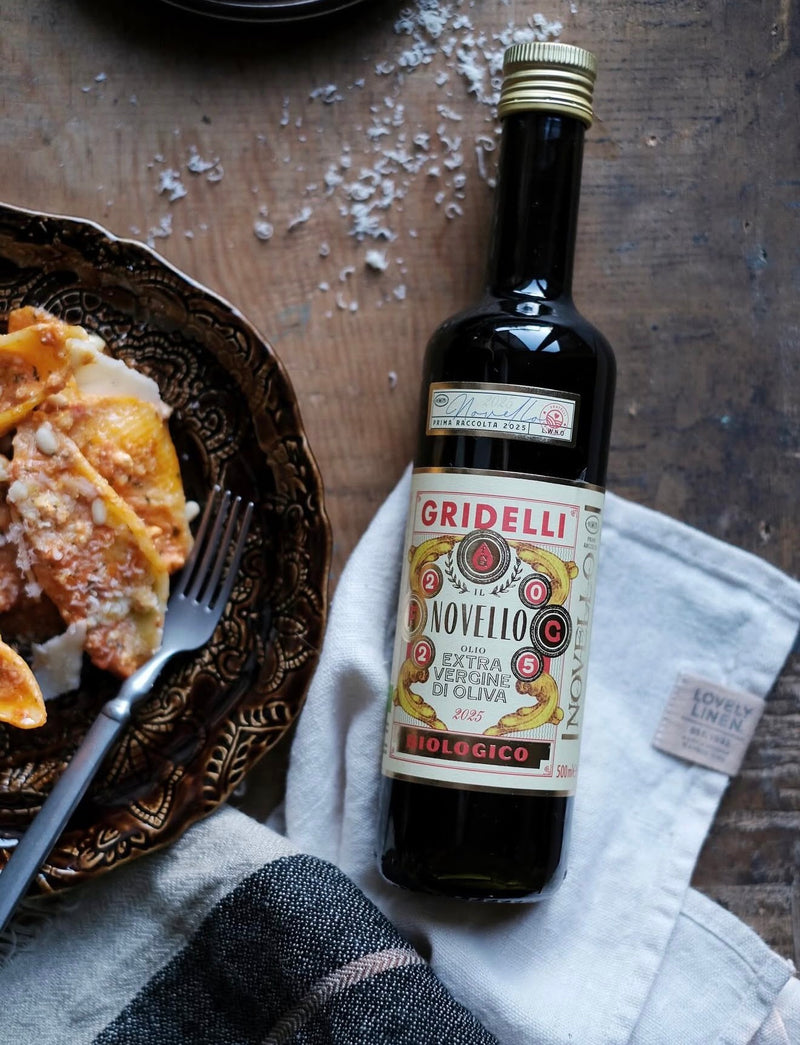 A bottle of Gridelli Novello extra virgine olive oil stands on a white cloth next to a plate of food on a rustic wooden table.