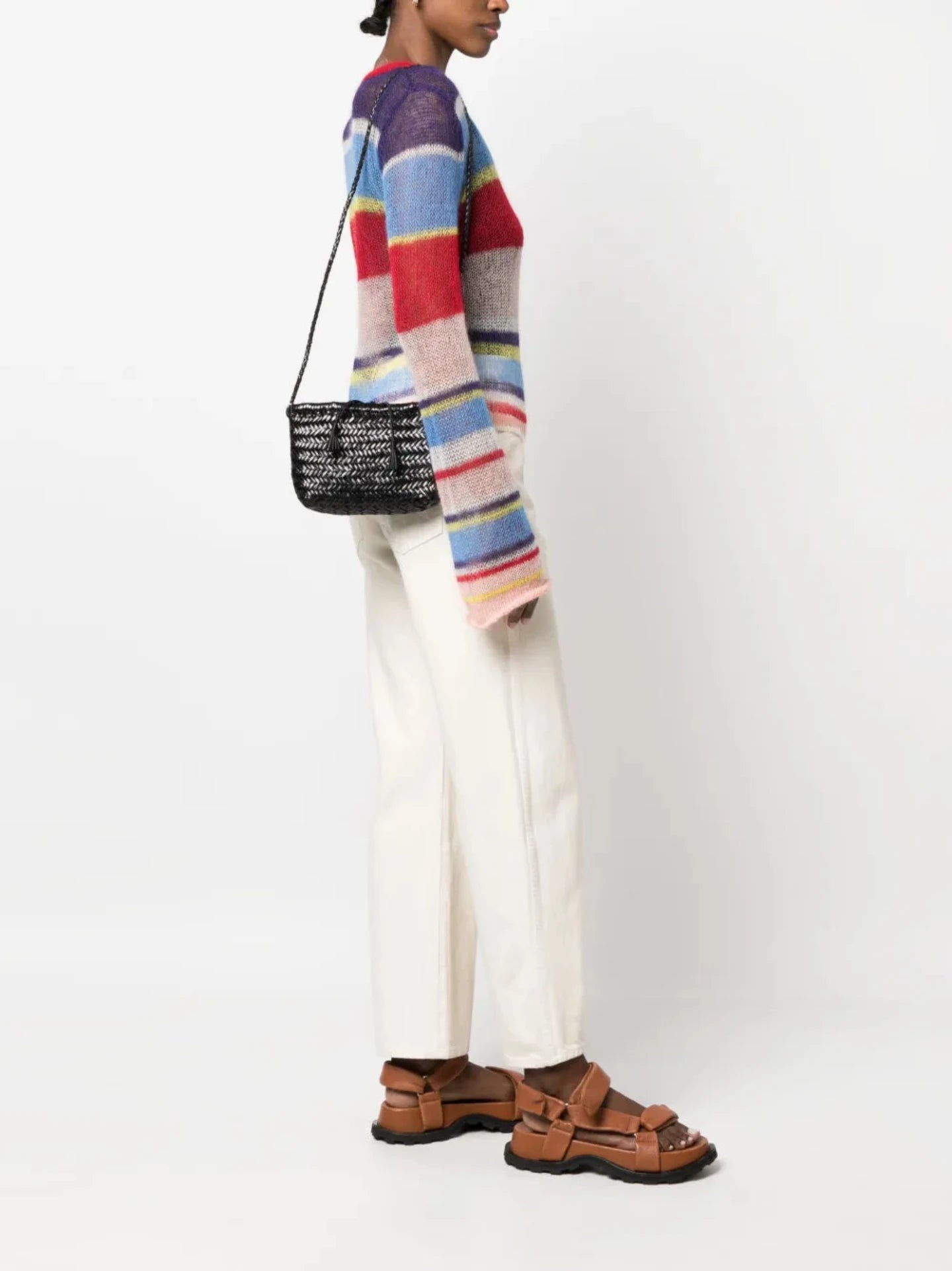 Person wearing a colorful striped sweater, white pants, and brown sandals, holding a black handbag on a white background.