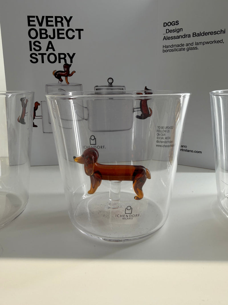 A clear glass tumbler featuring a small amber-brown glass dachshund figurine inside, displayed on a white surface with a white background and other glassware partially visible.