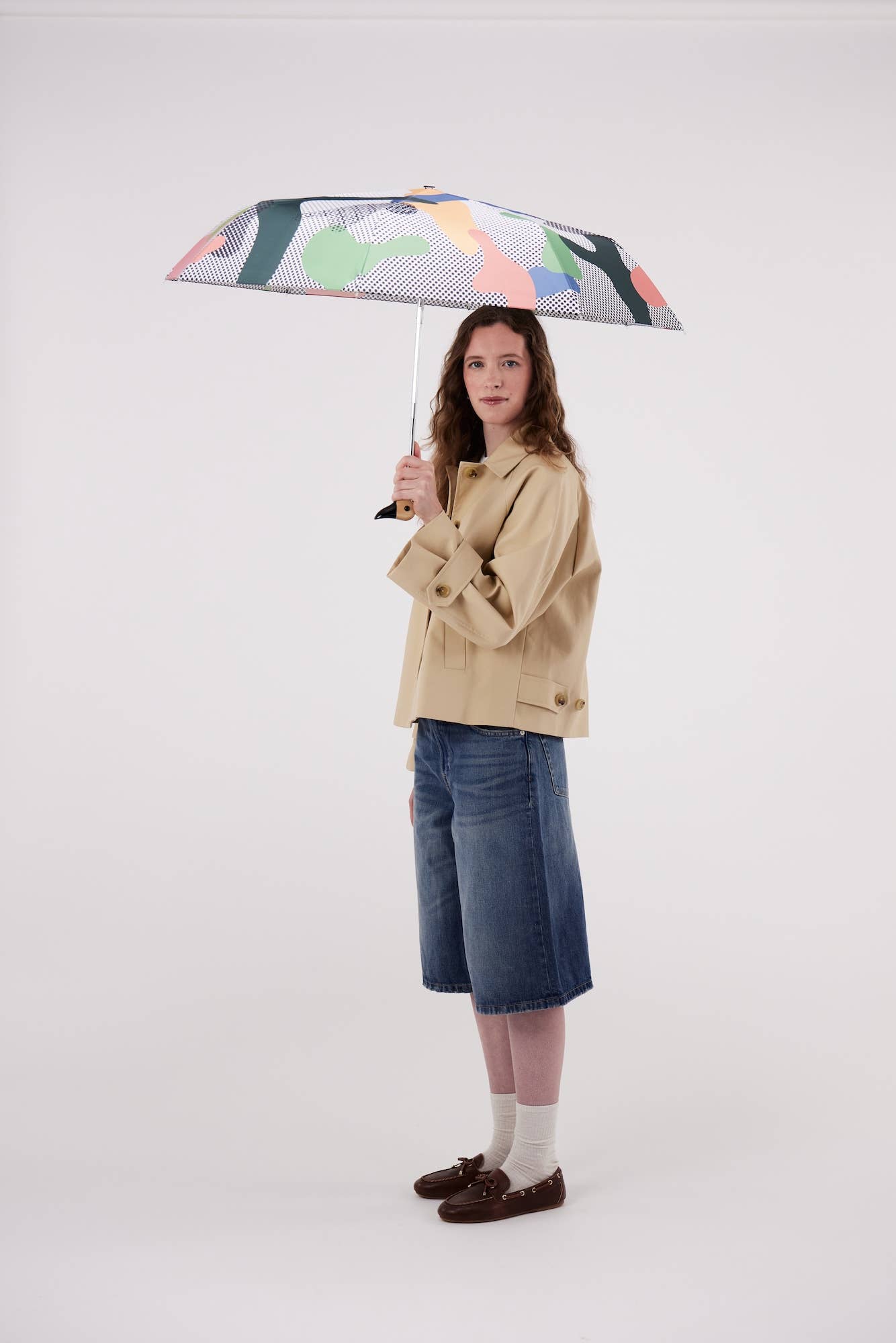 A model stands holding a colorful abstract-patterned umbrella, wearing a beige trench coat, blue denim shorts, white socks, and brown loafers against a plain light gray studio background.