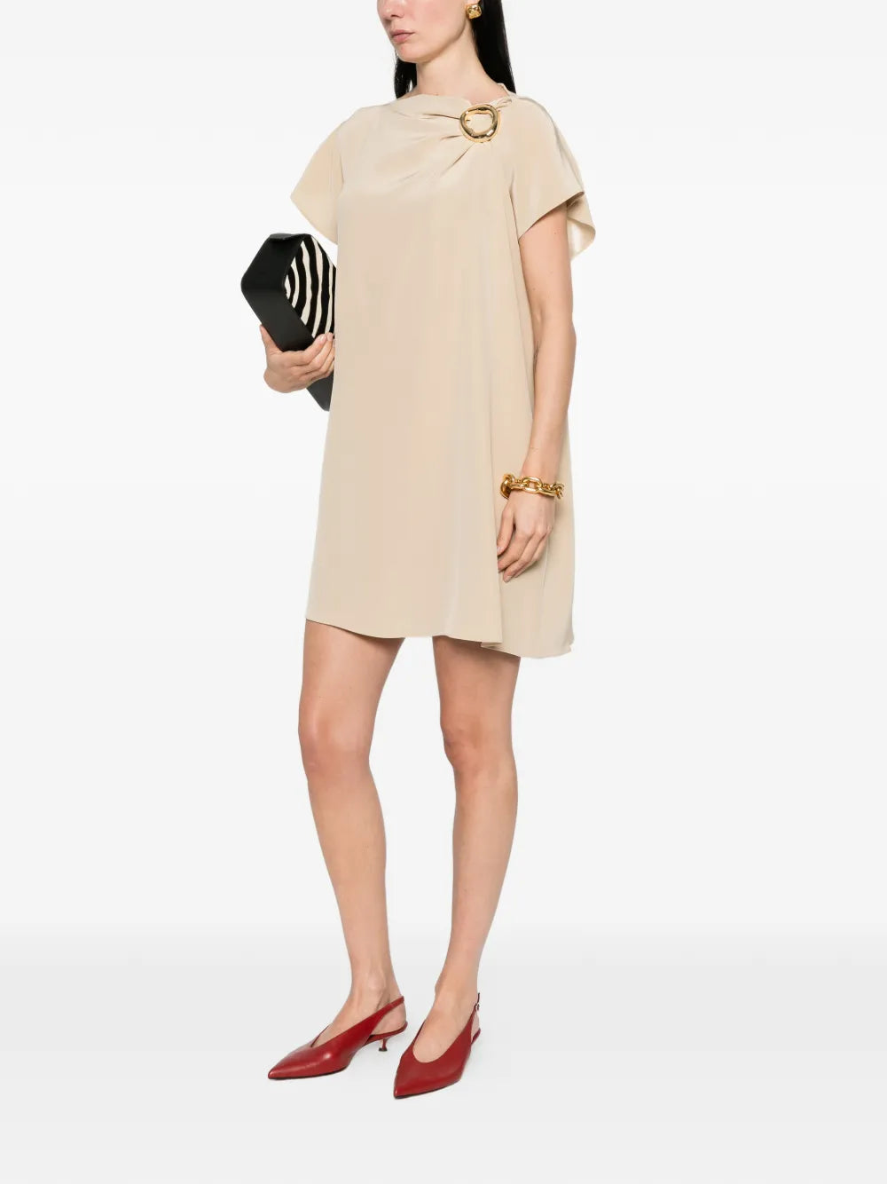 Woman wearing a beige dress with a gold belt, holding a black clutch on a white background