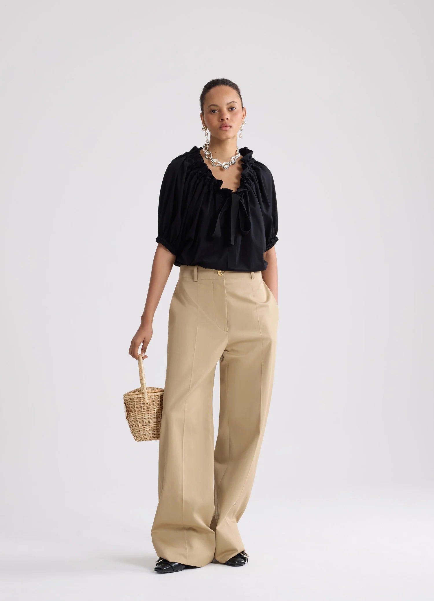 Woman wearing a black blouse and beige pants holding a woven bag on a white background