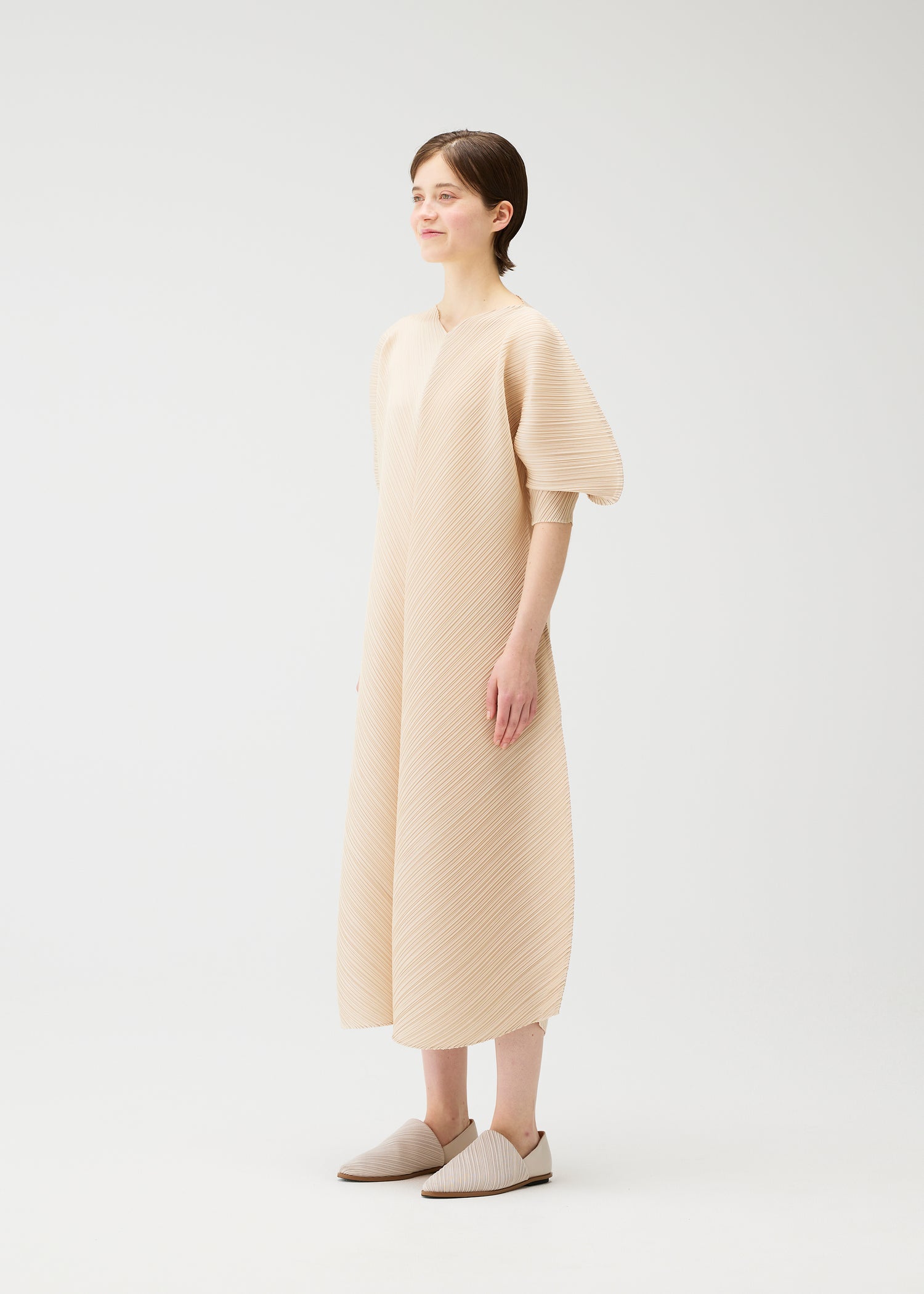 Model wearing a long beige short-sleeved dress from diagonally pleated fabric typical for the Pleats Please Issey Miyake on a white background