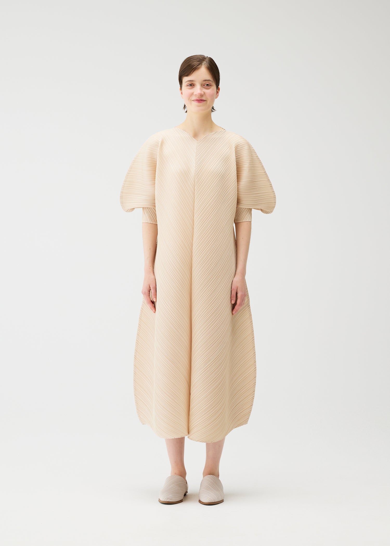 Model wearing a pale beige pleated dress with short bell-shaped balloon sleeves that are cut narrow standing against a white studio background