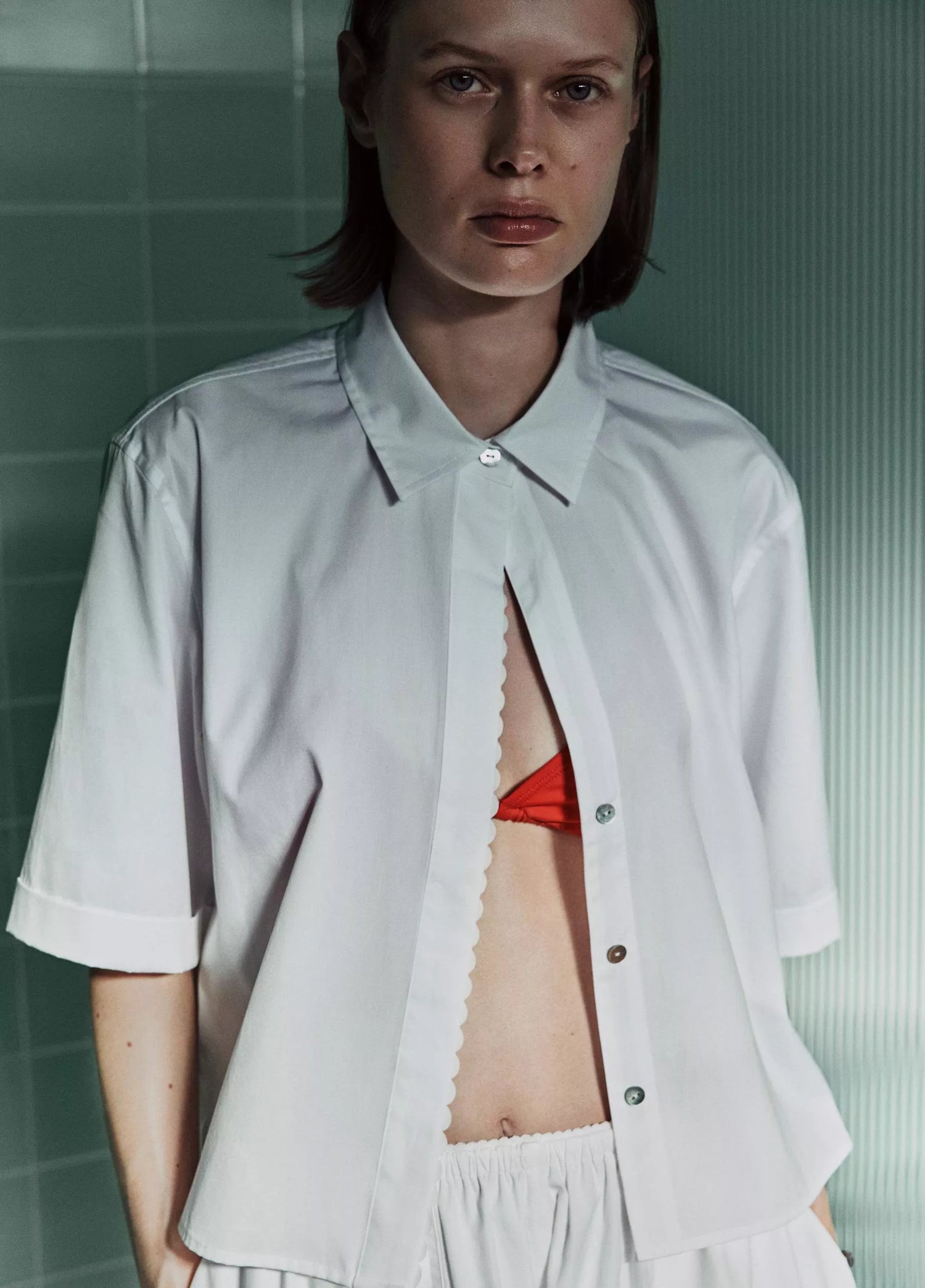 Person wearing a white shirt with a cut-out design, standing against a green tiled wall.