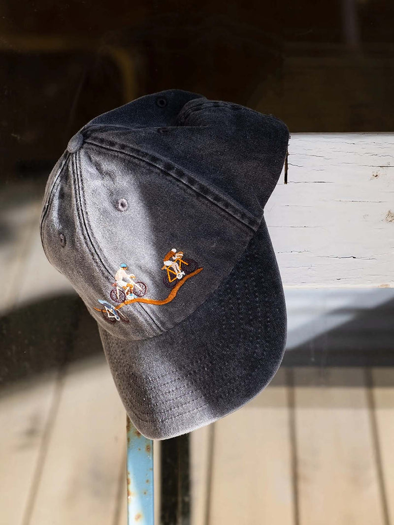 A washed black baseball cap with embroidered mountain biking figures, resting on a weathered white wooden surface.
