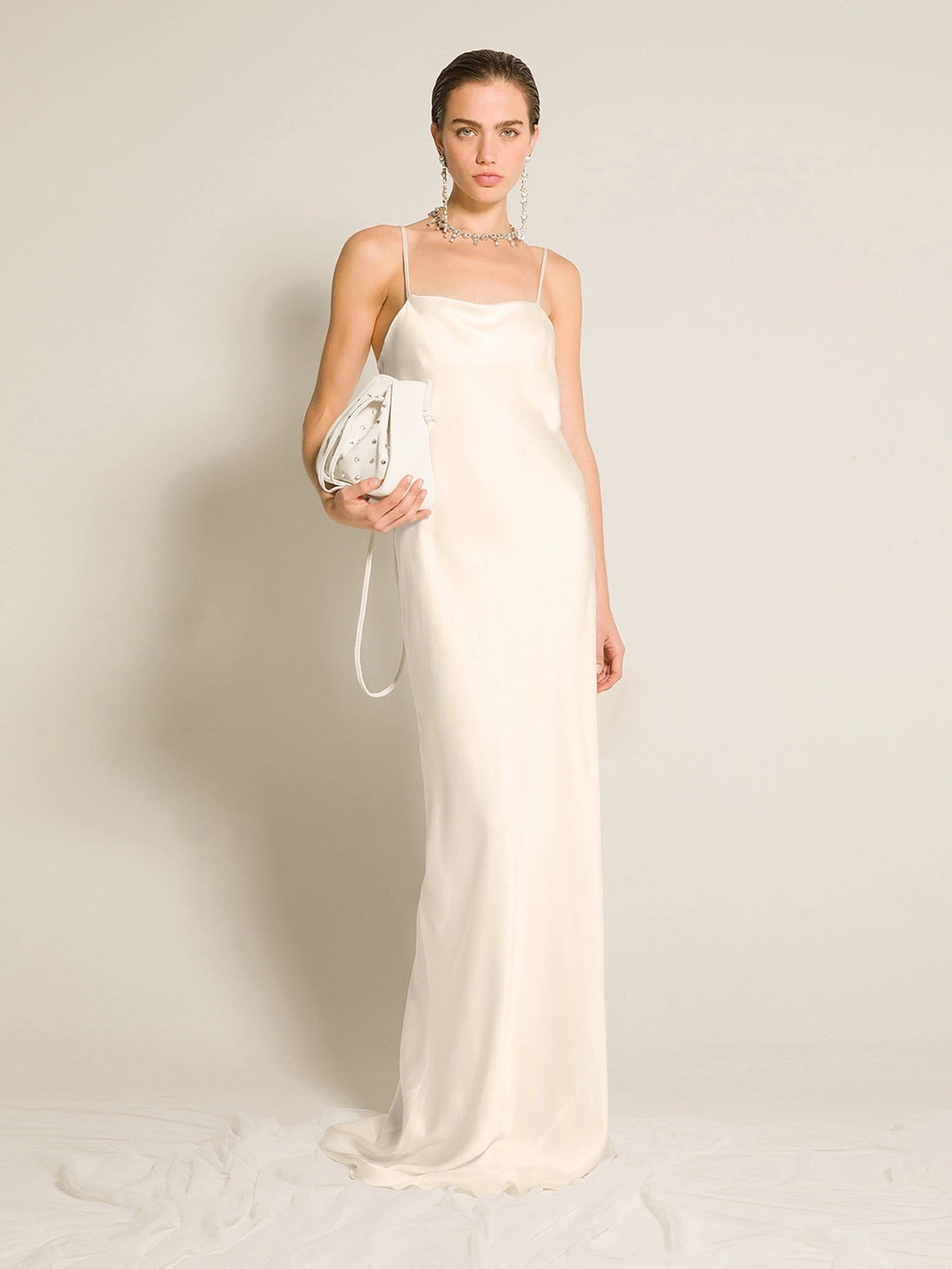 Woman in a long white dress holding a clutch against a plain background
