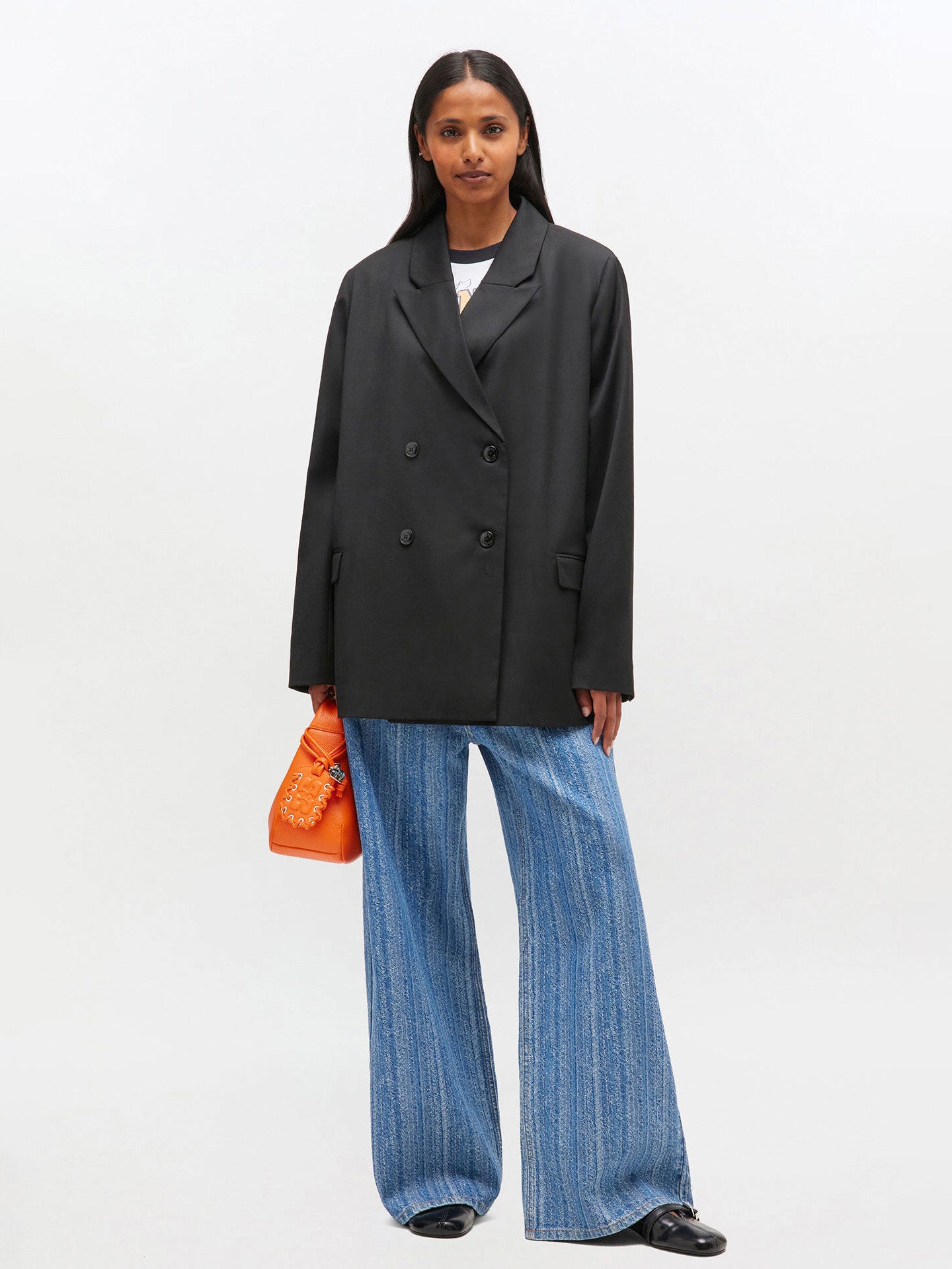 Woman wearing a black blazer and blue jeans with an orange bag on a white background