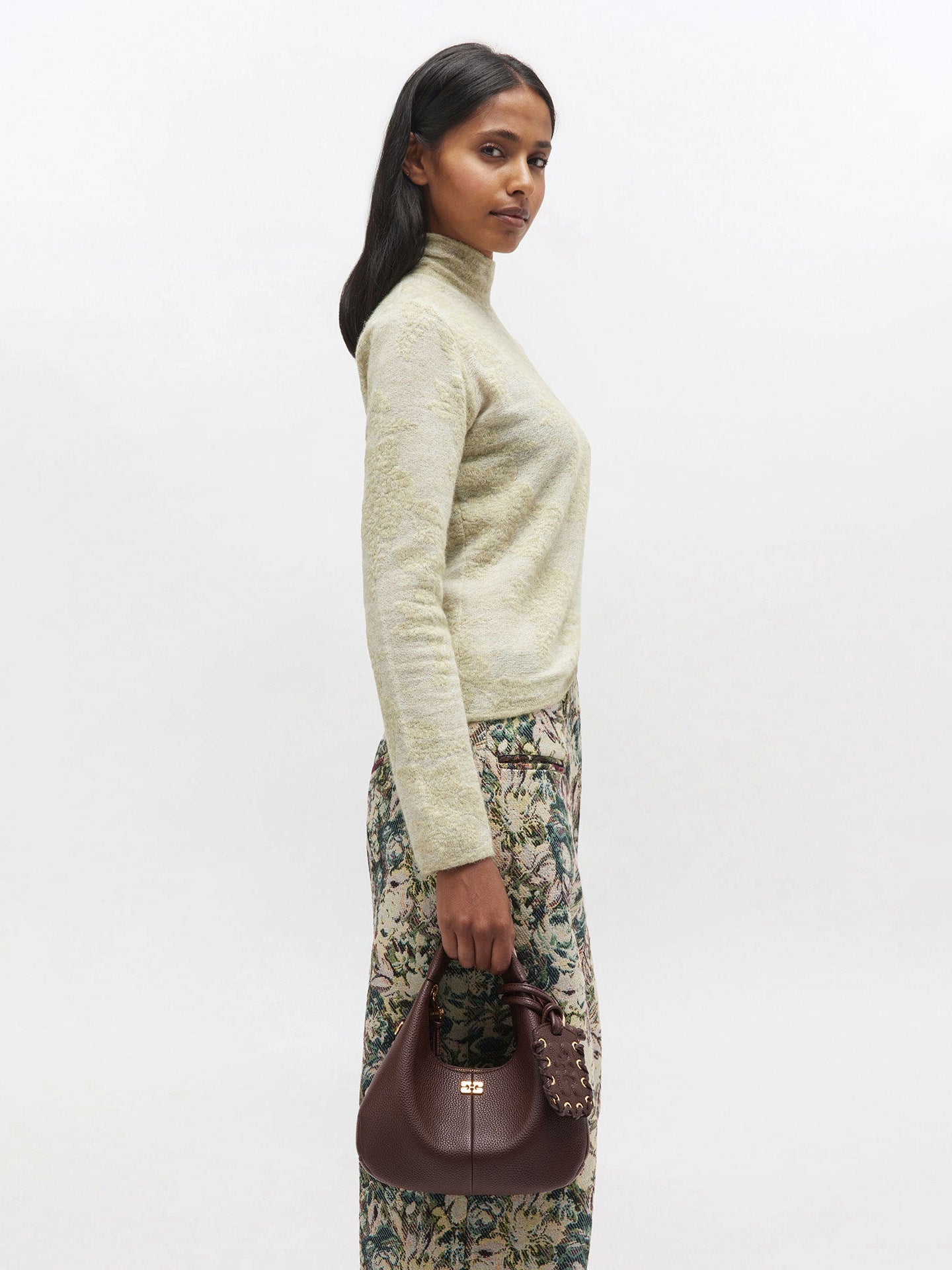 Woman wearing a cream sweater and floral skirt holding a brown handbag on a white background