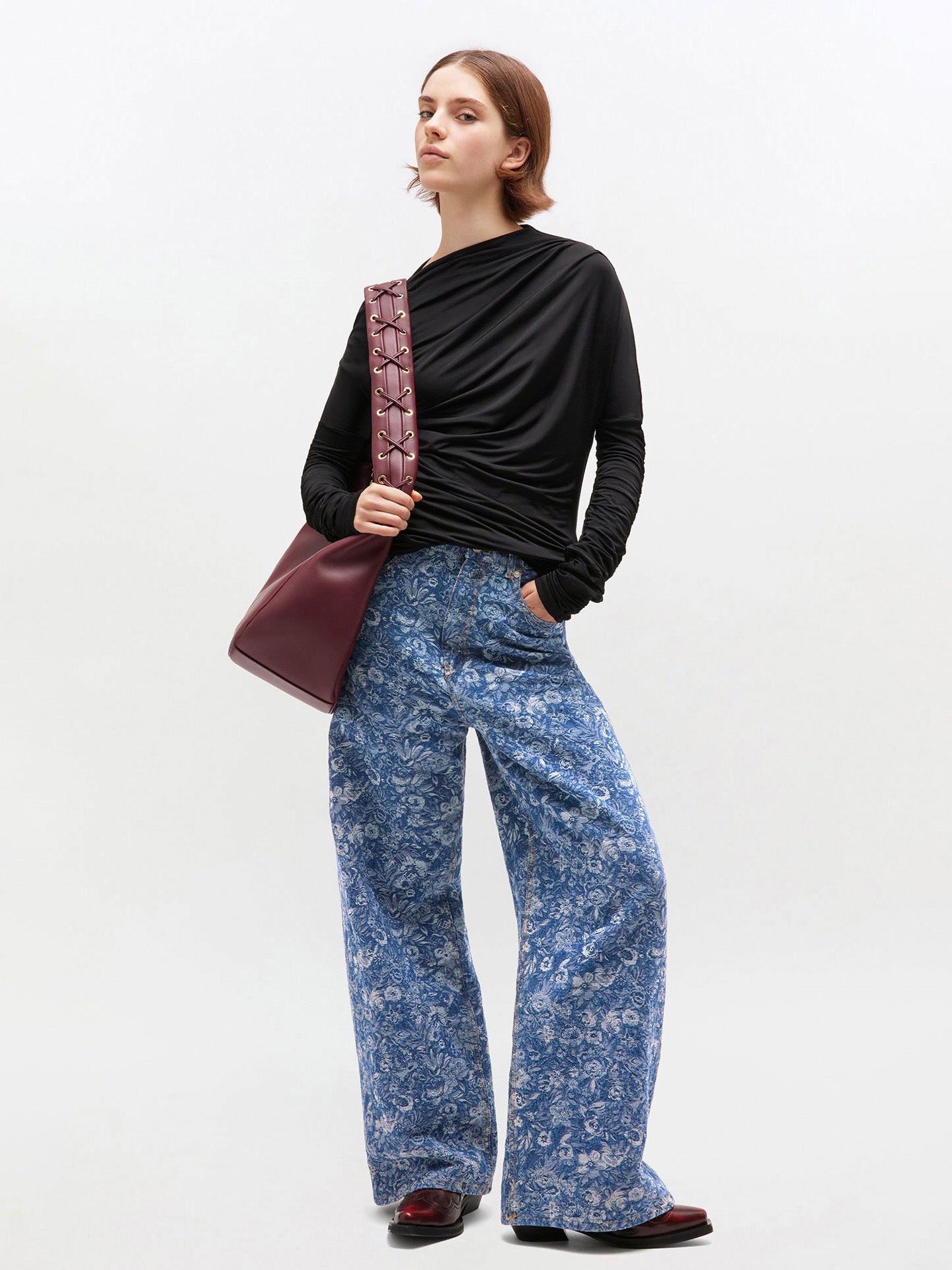 Woman wearing a black top and blue floral pants with a maroon bag on a white background