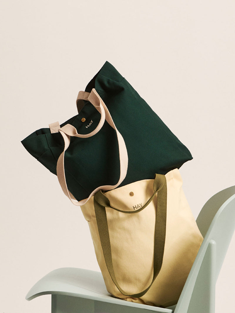 Two canvas tote bags from the brand HAY, one in dark forest green and one in beige, stacked on a light gray chair against a warm cream background.