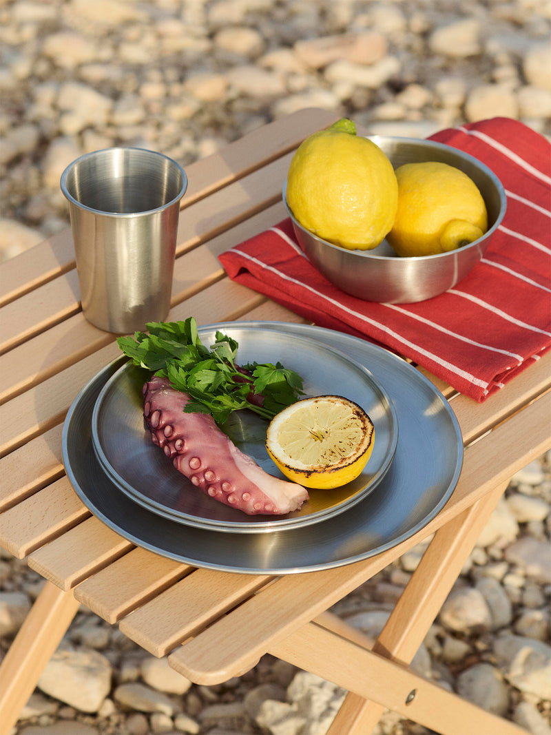 Outdoor setting with 3 minimalist stainless steel plates, one large dinner plate and 2 smaller side or salad plates stacked with calamari, grilled half lemon and fresh herbs, a similar style stainless steel bowl with 2 fresh lemons, and a matching large metal cup on a wooden table along with a red striped kitchen towel.