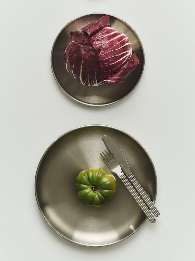 Two round brushed-metal plates on a light gray background. The smaller top plate holds a head of red cabbage, and the larger bottom is a dinner plate holding a green tomato with a fork and knife.