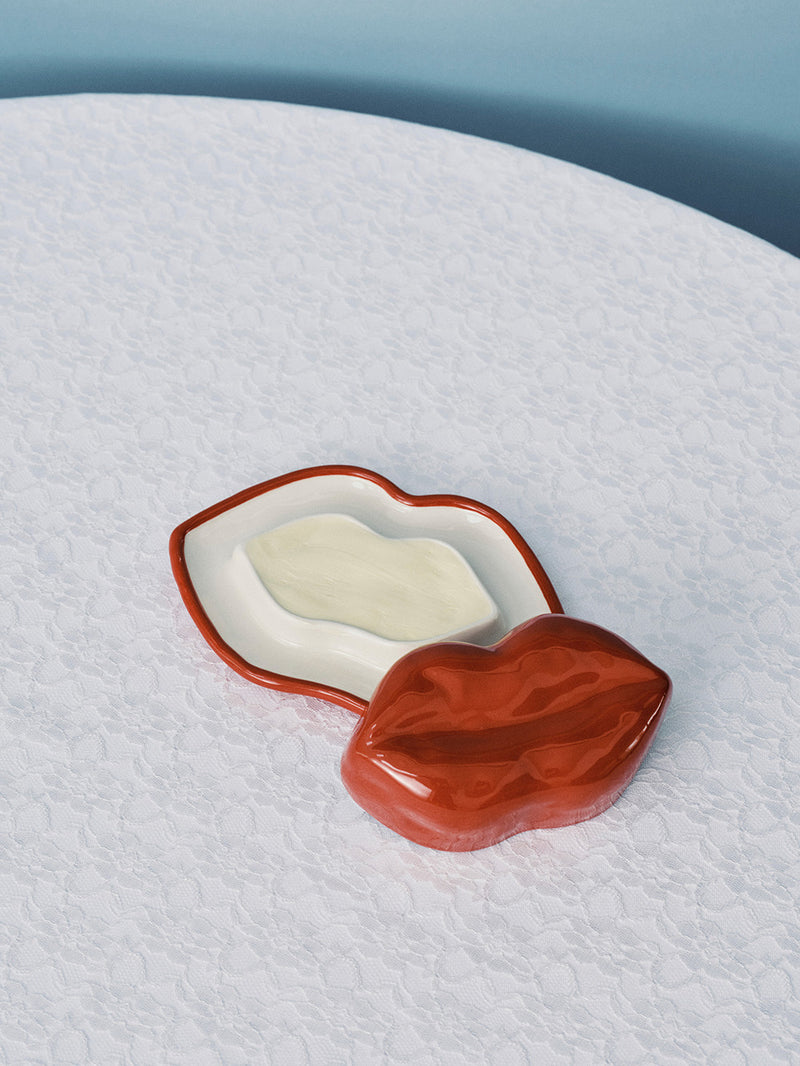 Red lips-shaped container with a white bar of soap on a textured white surface.